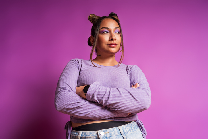 Studio portrait of a stylish young woman crossing her arms, conveying confidence and strength against a vibrant purple backdrop